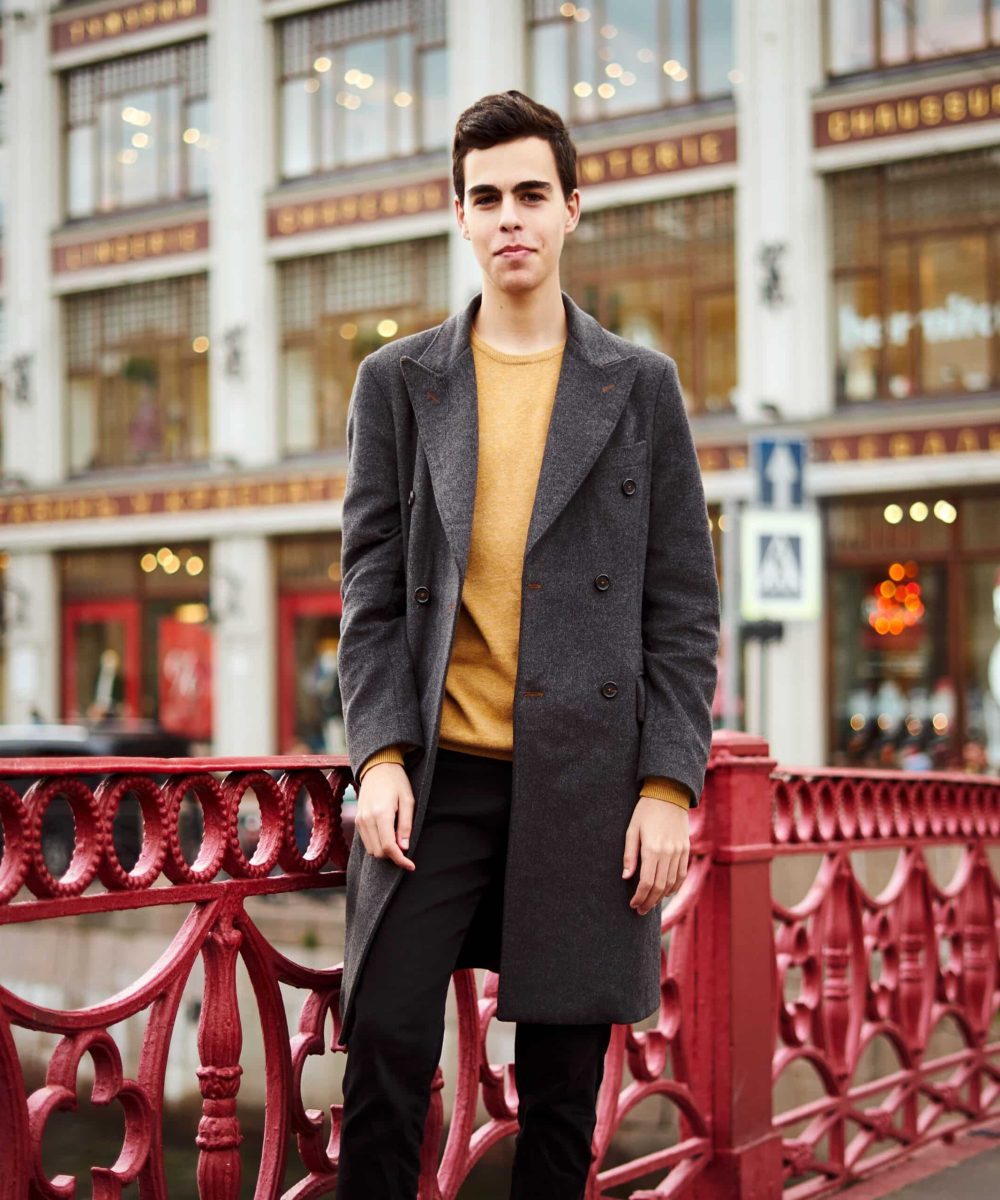 Handsome stylish fashionable man, brunette in elegant gray coat, stands on the street in historical center of St. Petersburg. Young man with dark hair, thick eyebrows.