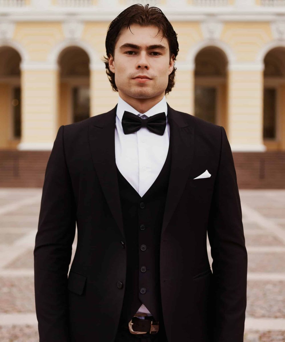 A handsome man wearing a black tuxedo with a bow tie stands outdoors. He poses in front of a building featuring arches and classical design.
